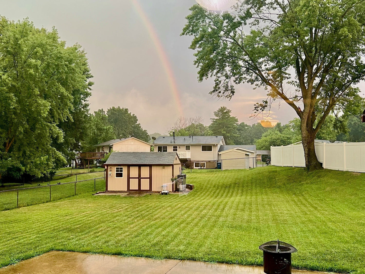 Gepflegter Garten mit Rasen, Gartenhaus, Bäumen, weißen Zäunen, Häusern im Hintergrund und Regenbogen.