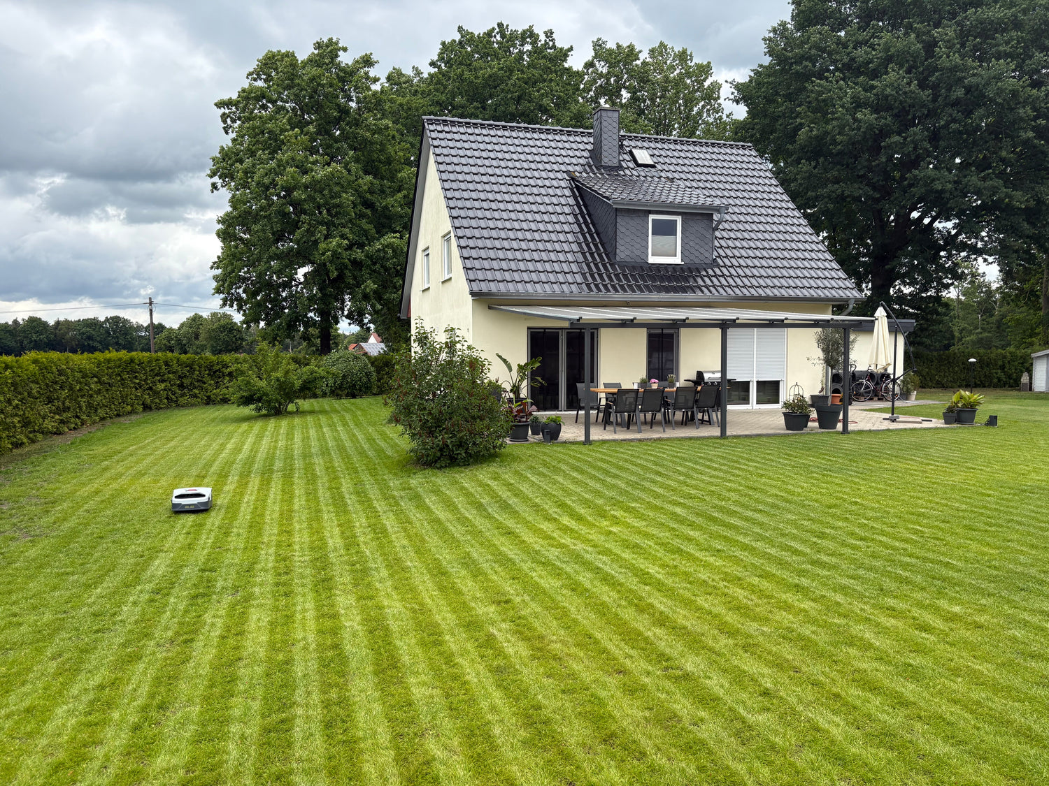 Ein gepflegtes Vorstadthaus mit dunklem Dach und Terrasse. Ein Mähroboter hat ein gestreiftes Muster in den grünen Rasen geschnitten.
